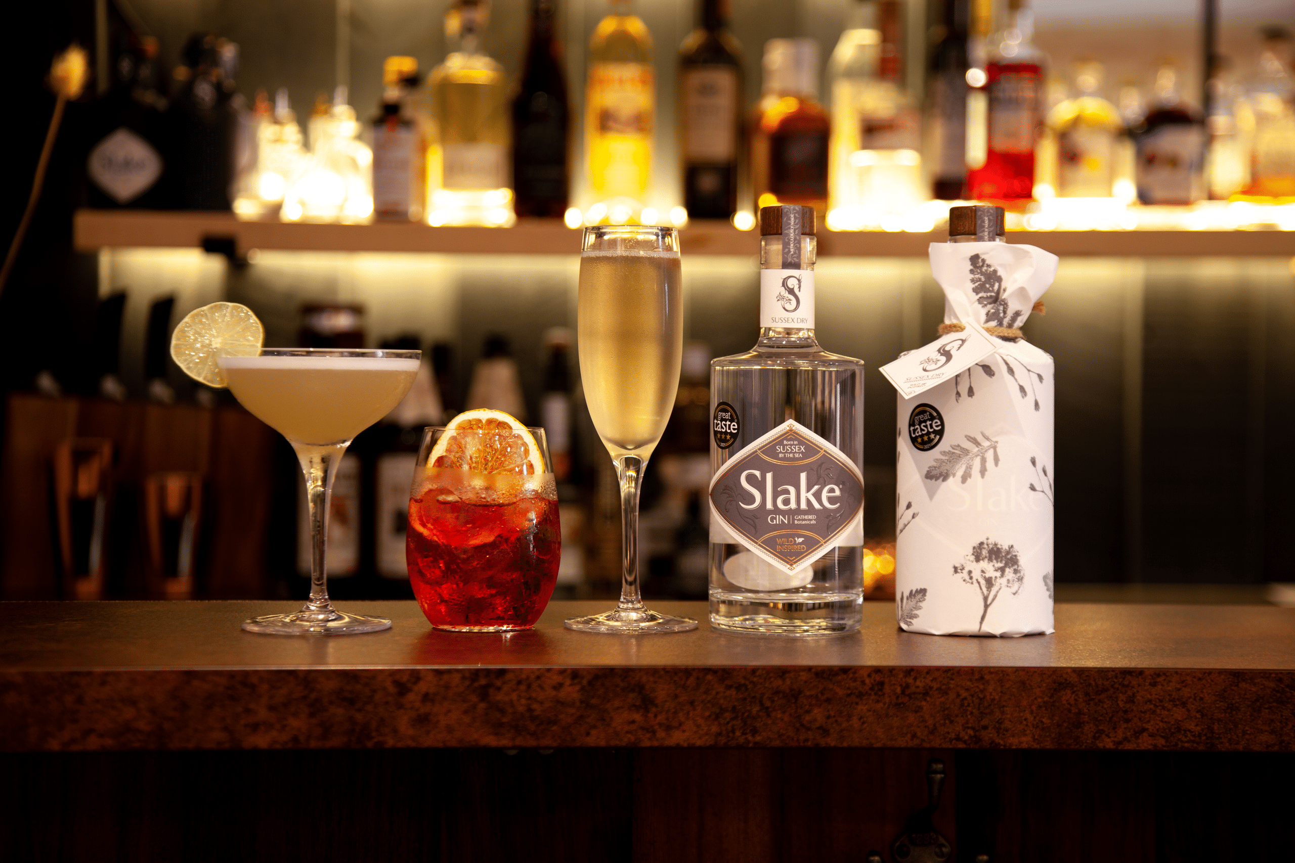 Slake Spirits Logo with copper botanicals and bottles of Sussex Dry Gin in industrial flint half recycled glass and Hedgerow Gin wrapped with aromatic botanical print Great Taste Golden Fork from the South East winner 2021