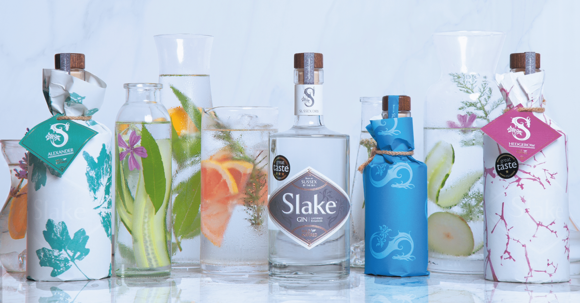 Slake Spirits Sussex Dry Gin Alexander Gin Hedgerow Gin and Wild Elixir Gin-based aperitif bottles on a white marble background with botanical drinks in glass flasks and scientific glassware.